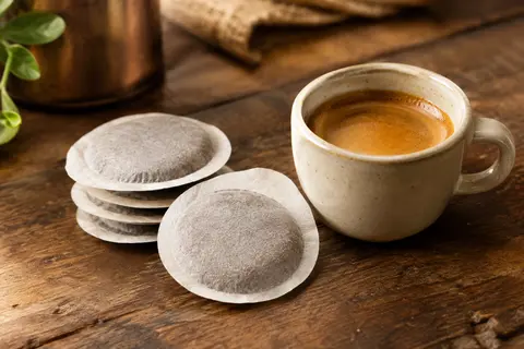 Dosettes de café ESE en papier filtre près d’une tasse d’espresso sur un comptoir en bois.