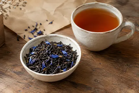 Thé noir Earl Grey en vrac avec fleurs de bleuet, présenté près d’une tasse ambrée sur du bois.