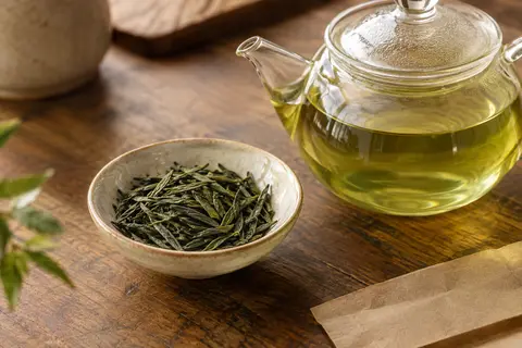 Feuilles longues de thé vert Sencha près d’une théière en verre contenant une infusion claire
