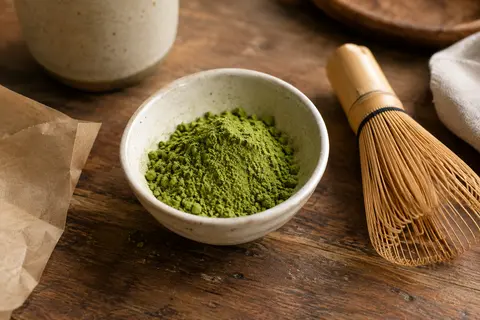 Poudre de matcha vert vif dans une coupelle en céramique, avec un fouet posé sur un comptoir en bois