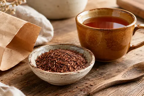 Rooibos en vrac brun rouge dans une coupelle, avec une tasse d’infusion ambrée sur un comptoir en bois