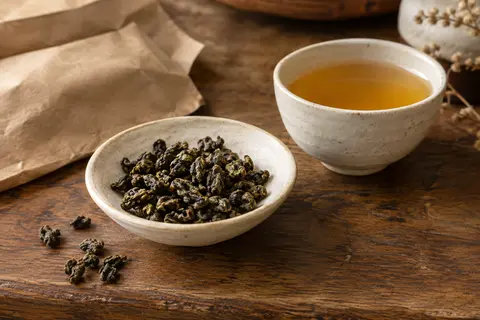 Feuilles roulées de thé oolong en vrac près d’une tasse d’infusion dorée sur un comptoir en bois.