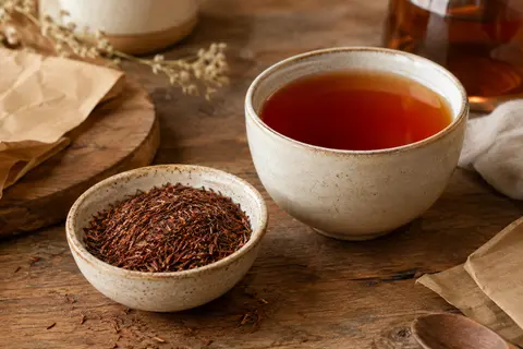 Feuilles de rooibos en vrac dans une coupelle, près d’une tasse d’infusion rouge orangé