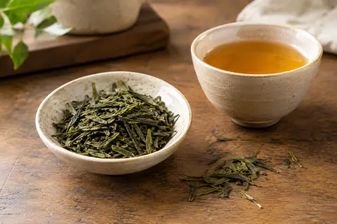 Feuilles de thé vert Sencha dans une coupelle en céramique, près d’une tasse d’infusion ambrée