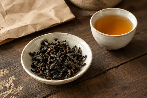 Feuilles torsadées de thé oolong Shui Xian dans une coupelle en céramique, près d’une tasse ambrée.