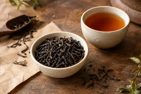 Feuilles de thé noir en vrac près d’une tasse ambrée sur un comptoir en bois