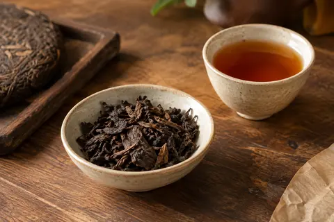 Feuilles de thé Pu-erh en vrac dans une coupelle en céramique, accompagnées d’une tasse ambrée